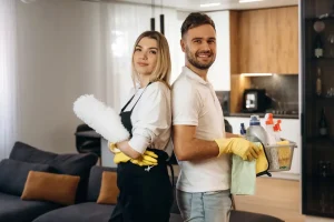 Two professional cleaners standing back to back while smiling confidently at the camera - they are holding various pieces of cleaning equipment